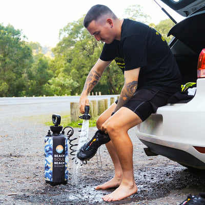 Photograph of a man with tattooed arms using a blue camouflage "Beach Soul Beach Shower" to rinse a black shoe while sitting barefoot on a white car's open trunk by a dirt road with lush green trees