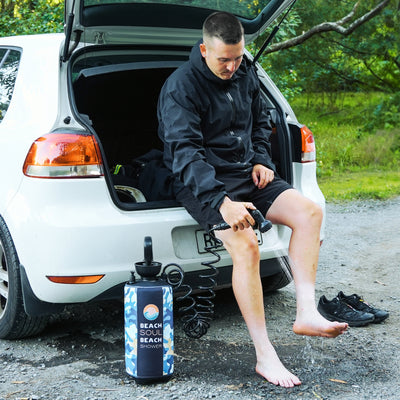 A man sitting on the open trunk of a white car uses a blue and white camouflage Beach Soul Beach Shower to rinse his bare feet on a gravel path near green foliage
