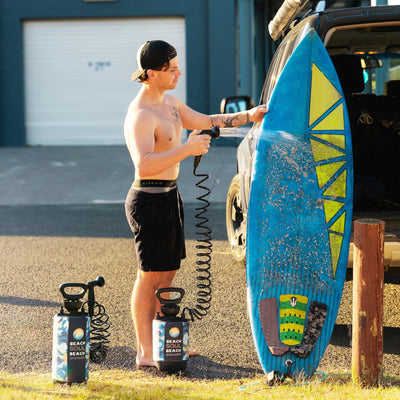 A shirtless man in a black cap and shorts rinses a sand-covered blue surfboard with yellow geometric patterns using a portable Beach Soul Beach Shower pump next to a dark SUV.