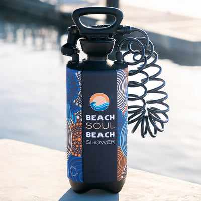 A Beach Soul Beach Shower portable pressure bottle featuring a dark blue neoprene sleeve with vibrant aboriginal-inspired patterns in blue, orange, and white, an orange sun and blue wave logo, and a black pump handle with a coiled hose, sits on a concrete surface near water under bright sunlight