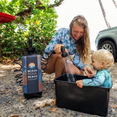 A smiling woman uses a portable Beach Soul Beach Shower with a vibrant blue and orange patterned cover to rinse a happy toddler in a black tub on a gravel path near palm trees and a light green SUV, the woman wearing a blue and white leaf-patterned rash guard and the child in a light blue marine-themed rash guard