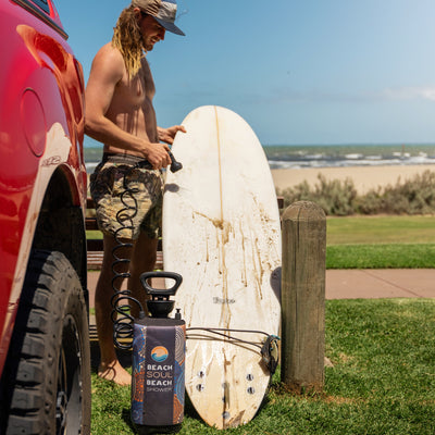 A smiling man with long blond hair and camouflage shorts uses a portable Beach Soul Beach Shower, featuring a colorful indigenous art pattern, to rinse a muddy white surfboard next to a red truck at a sunny beach