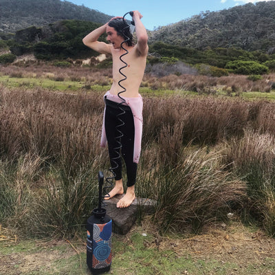 Photograph of a man in a pink wetsuit top and black leggings showering with water spraying from a portable outdoor shower featuring an Indigenous art-inspired patterned base in a reedy wilderness landscape with forested hills