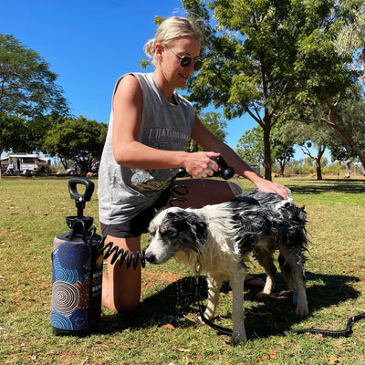 Photograph of a blonde woman in sunglasses washing a wet black and white dog in a sunny grassy campsite with a blue portable shower featuring an Indigenous-style dotted pattern and a white caravan in the distance