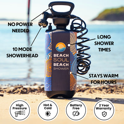 Product photography of the portable Beach Soul Beach Shower, a black pressure-activated unit with a colorful blue and orange patterned thermal sleeve and a 10-mode showerhead, set on a bright sandy beach beside the calm ocean with a distant landmass