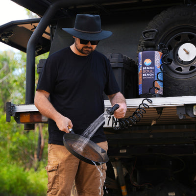 A man wearing a black fedora and sunglasses washes a pan with a portable shower featuring a colorful patterned Beach Soul sleeve and coiled black hose beside an off-road vehicle