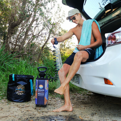 A young man wearing a cap, sunglasses, and turquoise towel sits in the open boot of a white car, rinsing his feet with a colorful "Beach Soul Beach Shower" next to a matching black bucket in a lush bushland setting
