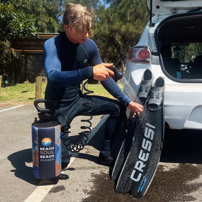 Photograph of a young man in a navy Billabong wetsuit cleaning black Cressi diving fins with a colorful Beach Soul Beach Shower portable shower next to a white car in a sunny outdoor parking area