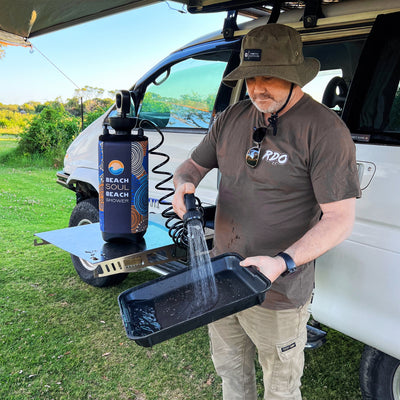 Photography of a man in an olive bucket hat using a portable Beach Soul Beach Shower with a blue and orange wave pattern to rinse a black tray, outdoors next to a white off-road vehicle with an extended awning
