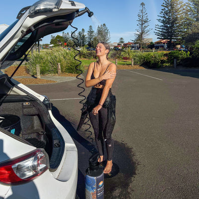 A joyful woman rinses off using a portable shower connected to a white car's open trunk in a sunny coastal parking lot, wearing a black bikini top and wetsuit bottoms, with tall Norfolk pines and a colorful Beach Soul Shower tank visible