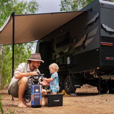 A man wearing a wide-brimmed hat showers a young child in a blue patterned romper in a black tub beside a dark grey Great Aussie Caravans Tribal Grove 216 caravan with a beige awning and a vibrant Beach Soul portable shower in an outdoor bushland setting