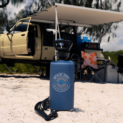 Blue "Beach Soul Beach Shower" portable shower on a sandy beach campsite with a yellow overland truck, extended awning, and an orange floral towel visible in the background