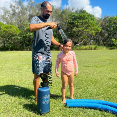 Photograph of a man in floral swim shorts rinsing a young girl with pink face paint and a pink long-sleeve swimsuit using a blue portable Beach Shower in a grassy outdoor area with blue pool noodles nearby