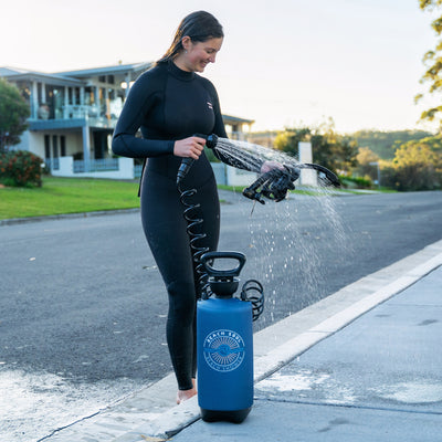 Photograph of a smiling woman in a black wetsuit with wet hair using a portable blue Beach Soul Beach Shower to rinse black diving gear on a suburban street at golden hour
