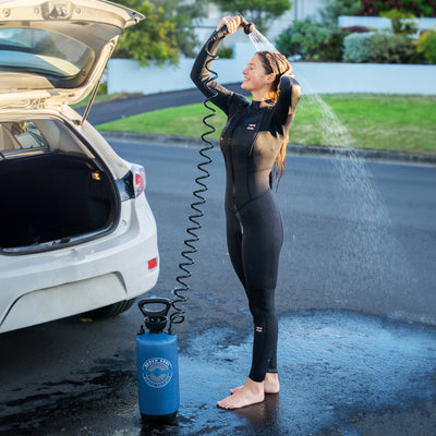 Photograph of a smiling woman in a black Billabong wetsuit rinsing her hair with a blue Beach Soul portable shower next to an open white hatchback car on a sunny asphalt street
