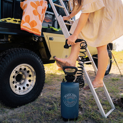 A person rinsing their sandy feet with a dark blue Beach Soul portable shower while standing on a ladder next to a beige off-road vehicle with an orange floral towel and rugged tire in a sandy outdoor setting