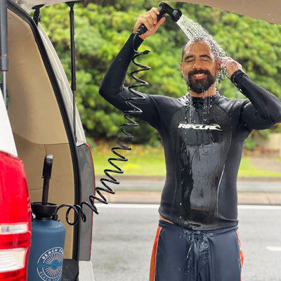 A smiling man in a black Rip Curl wetsuit and orange-striped board shorts rinses off with a coiled hose portable shower system from a blue Beach Shower bottle at the rear of a white van against a lush green background