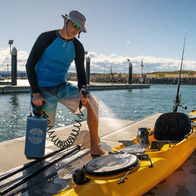 A man in a grey hat and green reflective sunglasses uses a blue Beach Soul Beach Shower portable sprayer to rinse his bright yellow fishing kayak on a sunny marina dock with sparkling water and a fishing rod in a holder
