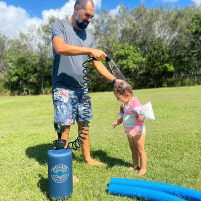 Bearded man in floral swim shorts rinsing a young child with wet hair, wearing a striped pink swimsuit and a "Frozen" arm float, using a blue Beach Soul portable shower on bright green grass with blue pool noodles under a sunny sky