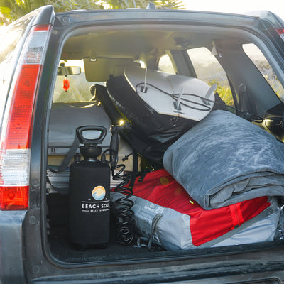 Photograph of an SUV trunk packed with beach and surf gear including a white surfboard, a black Beach Soul portable shower, and a red bag under sunny skies