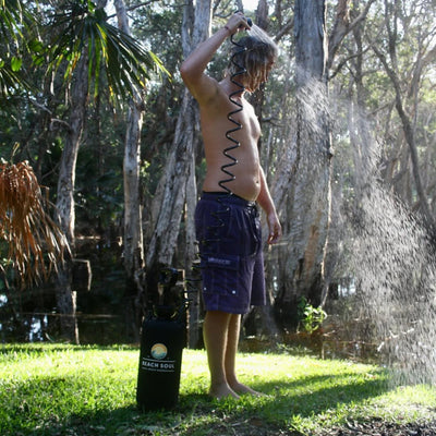 Photography a shirtless man with wet blonde hair showers outdoors in a lush natural setting using a black coiled hose from a Beach Soul portable shower system, wearing dark purple Billabong board shorts, standing barefoot on vibrant green grass with sunlit water spraying to his right