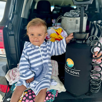 Adorable toddler with sandy cheeks in a blue and white striped robe sits in an open car trunk, playfully holding a black Beach Soul portable beach shower with its vibrant wave logo and coiled hose, surrounded by colorful beach towels
