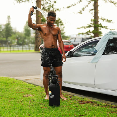A shirtless wet man showers next to a white car on a grassy verge, holding a black portable shower head, with a Beach Soul branded tank on the grass and a light blue towel draped over the car door