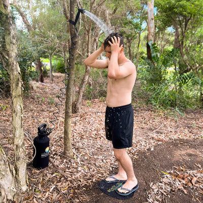 A man with wet hair stands on an oval black 'BEACH SO' branded mat showering outdoors from a portable black coiled hose shower system attached to a tree in a lush green forest wearing black board shorts and flip-flops