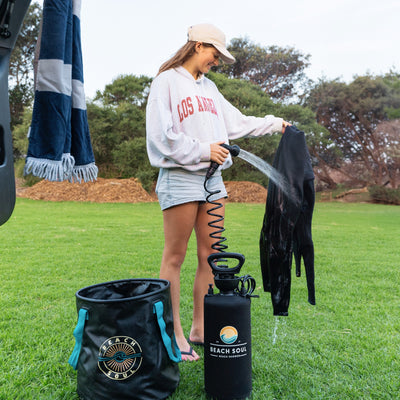 A young woman in a beige Los Angeles hoodie and denim shorts uses a black Beach Soul portable shower to rinse a wetsuit on a green grassy field, with a matching Beach Soul collapsible bucket and a blue and white striped towel nearby