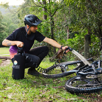 Photograph of a man wearing a black helmet and black and purple shirt cleaning his mud-splattered black mountain bike with a black Beach Soul portable shower while crouching on green grass in a sun-dappled forest