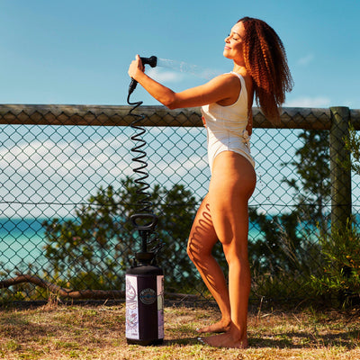 Photograph of a joyful woman with vibrant red curly hair in a white one-piece swimsuit showering herself with a stream of sparkling water from a black and pink portable shower featuring "Beach Soul" art, set against a chain-link fence overlooking a clear blue ocean