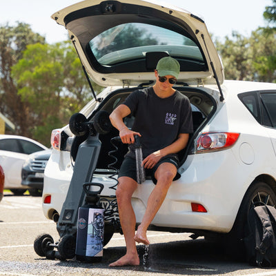 A young man wearing a green cap and a "GOOD DAY TO EXPLORE" t-shirt sits on the open trunk of a white car, rinsing his bare feet with a black portable shower featuring a "BEACH SOUL" skeleton illustration, next to an electric skateboard in an outdoor parking lot
