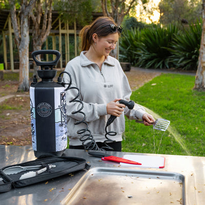 A smiling woman with sunglasses on her head wearing a grey "Surf" sweatshirt rinses a metal spatula with a coiled hose from a portable "Beach Soul Beach Shower" bottle on a stainless steel outdoor sink alongside a red knife and white cutting board in a sunny park setting