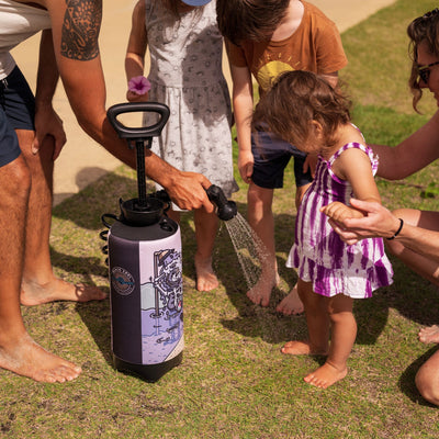 A family rinses sandy bare feet on a sunny grassy lawn with a portable black shower featuring a skeleton surfing graphic, highlighting a man's tribal arm tattoo and a child in a purple tie-dye dress