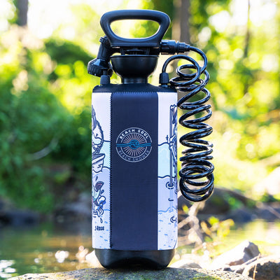 Product image of a black portable Beach Soul pressure shower with a coiled hose and trigger nozzle, adorned with a navy blue and white neoprene sleeve featuring artwork by J Ross including a central sun and wave logo, a shark, and a skeleton figure, resting on a mossy rock beside a sunlit stream in a vibrant green outdoor setting