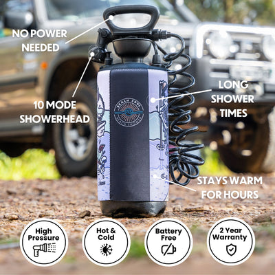 Photograph of a portable Beach Soul Beach Shower unit with a purple and black neoprene sleeve featuring a sun and waves logo and beach-themed cartoon graphics, on dirt ground with an off-road vehicle in the background, highlighting its battery-free design and 10-mode showerhead
