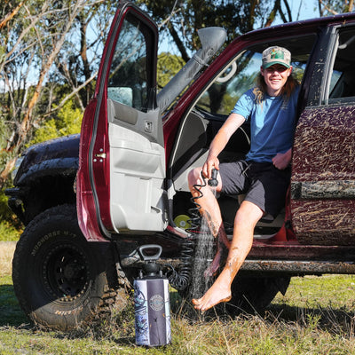 Photograph of a smiling man in a fishing cap showering his muddy legs with a J Ross portable Beach Shower while sitting in a mud-splattered burgundy 4x4 with a snorkel in a sunny natural outdoor setting