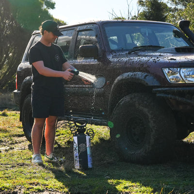 A man in a dark cap and shorts cleans a mud-splattered burgundy Nissan Patrol SUV with a portable spray washer in a sunny grassy field with trees