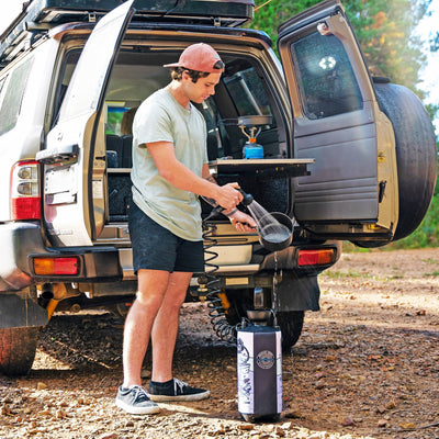 Photography of a man in a peach cap and light green shirt rinsing a pot with a portable shower attached to a silver Nissan Patrol SUV, its open tailgate kitchen featuring a blue gas stove and a black and pink Beach Soul water tank in a sunny Australian campsite