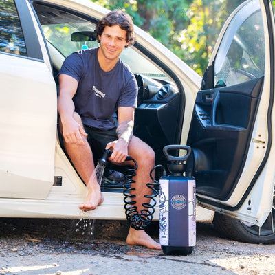 Photograph of a smiling man in a dark gray Billabong t-shirt sitting in an open white car door outdoors rinsing his bare feet with a portable Beach Soul Beach Shower featuring a coiled black hose