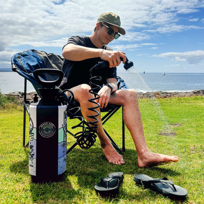 A man in a "HAPPY" cap and sunglasses showers his leg with a "BEACH SOUL" portable shower on vibrant green grass beside a rocky coastline with sailboats on the blue ocean