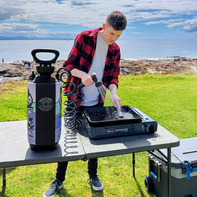 A man in a red and black plaid shirt uses a portable Beach Soul shower with a colorful wave design to wash a tray on an All 4 Adventure cooker at a Coleman table near a rocky coastline with sailboats on the distant ocean