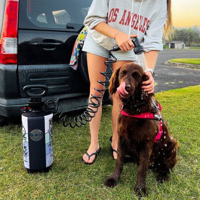 A person in a gray Los Angeles hoodie showers a brown dog with a pink harness licking its nose using a black Beach Soul portable shower unit next to a black SUV on a grassy verge with a distant white shed