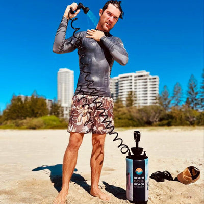 Photograph of a man in a wet grey rash guard and brown floral swim shorts using a portable black Beach Soul shower with a blue water stream to rinse off on a sunny sandy beach, tall white high-rises visible under a clear blue sky, with a brown cap and black flip-flops on the sand nearby