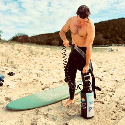 Bare-chested man in black wetsuit bottoms rinsing off with a portable black Beach Soul Beach Shower next to a light teal surfboard on a golden sandy beach under a partly cloudy sky