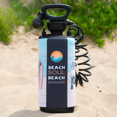 Photograph of a portable beach shower with a black handle, sandy nozzle, and coiled hose standing on golden sand, featuring a light blue neoprene sleeve with a Beach Soul Beach Shower logo, a pink surfboard illustration, and a white van graphic against a blurred green foliage background