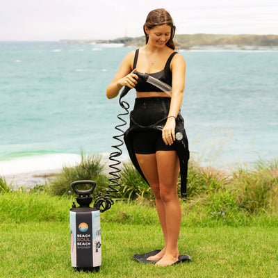 A smiling young woman in black wetsuit shorts rinses her arm with a portable Beach Soul Beach Shower unit featuring a colorful surf graphic, standing barefoot on green grass overlooking a turquoise ocean with a distant headland