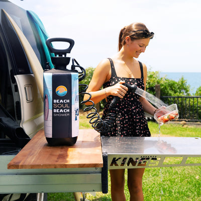 A young woman in a floral dress washes fresh strawberries using a portable Beach Soul Beach Shower system on a Kings Adventure outdoor table next to a van with surfboards overlooking the ocean