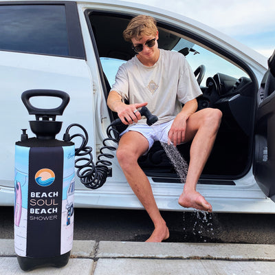 Photography of a young man in sunglasses sitting in a white car doorway washing his bare foot with a portable "Beach Soul Beach Shower" pump featuring colorful surfboards and a camper van graphic on its insulated sleeve