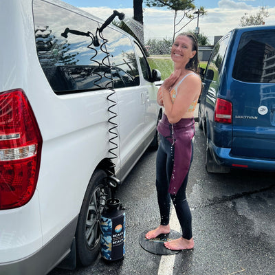 Photograph of a smiling woman using a portable Beach Soul Beach Shower attached to a white van, wearing a colorful floral bikini top and black wetsuit with a burgundy top pulled down, standing on a black textured mat in a parking lot with a blue Multivan and distant ocean visible